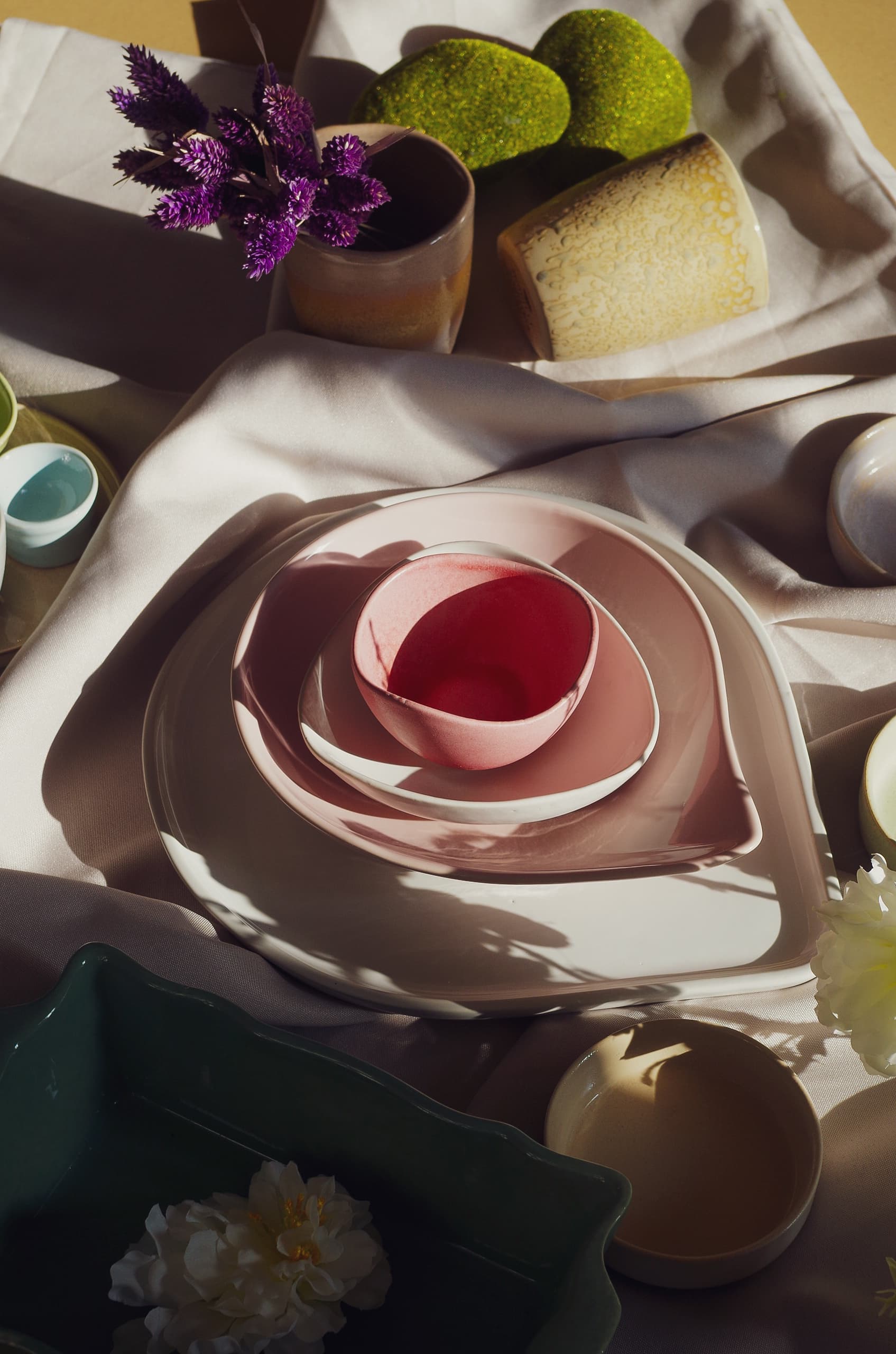 Mise en scène lifestyle d'une collection de céramiques aux tons roses, verts et bruns, assiettes et bols empilés sur tissu blanc avec fleurs séchées et lumière naturelle