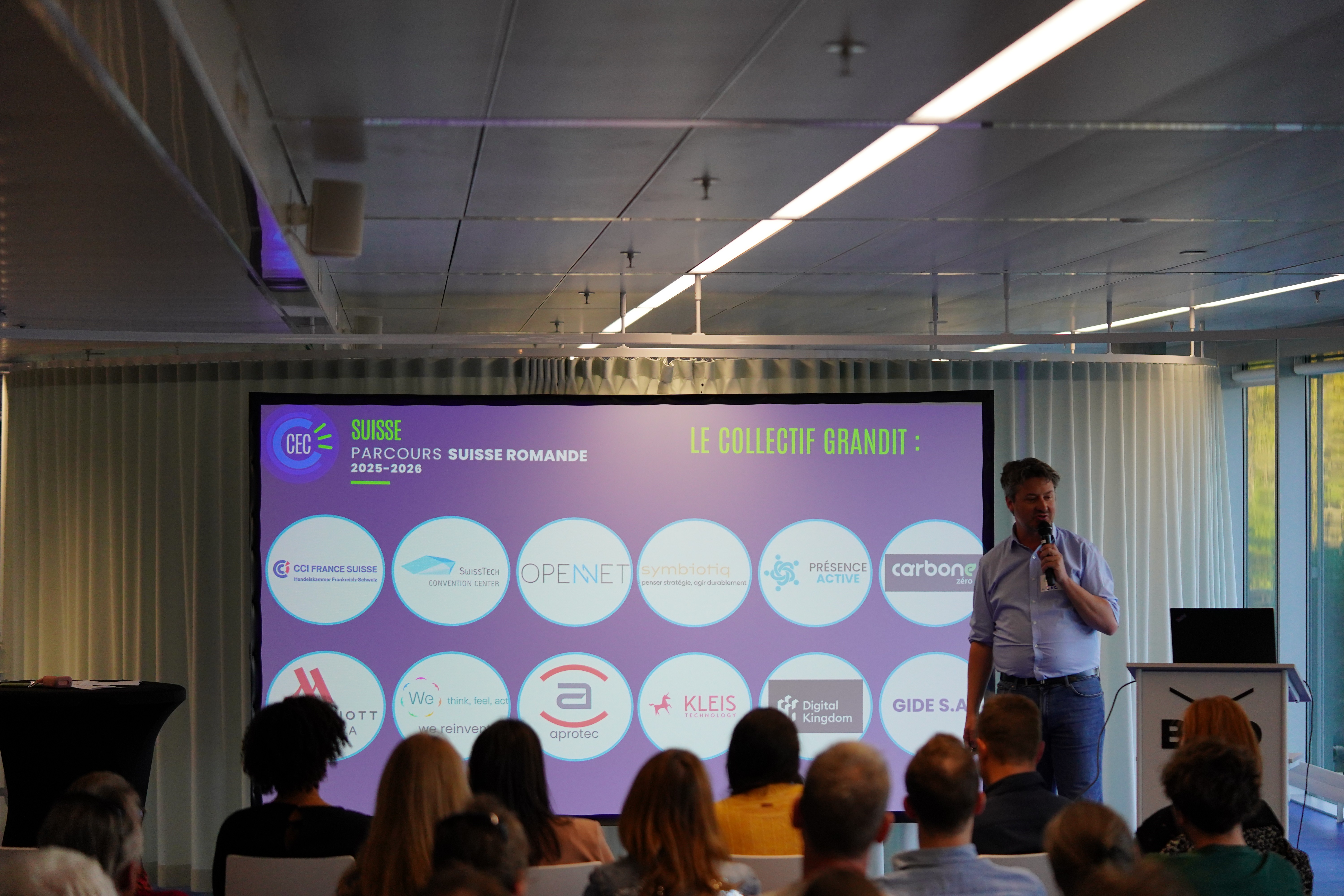 A smiling young man with crossed arms, wearing a plaid shirt and white t-shirt, poses against a dark background.