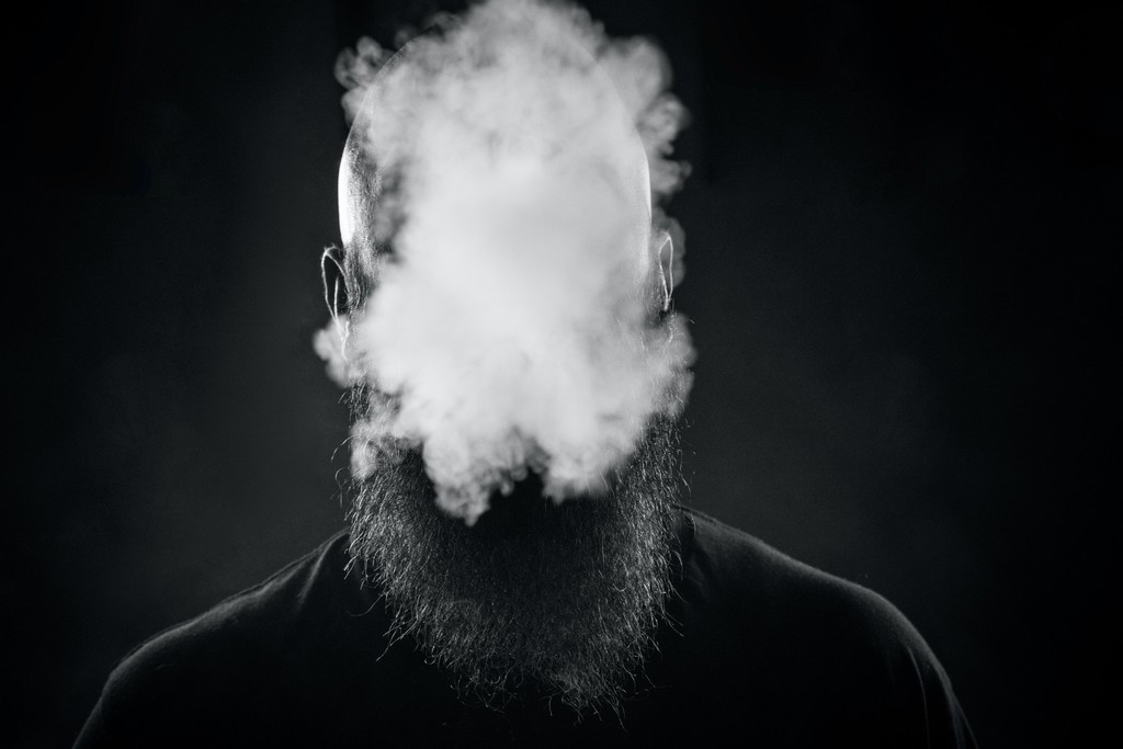 close up portrait in black n white showing a bald man with a huge beard covering his face with smoke.