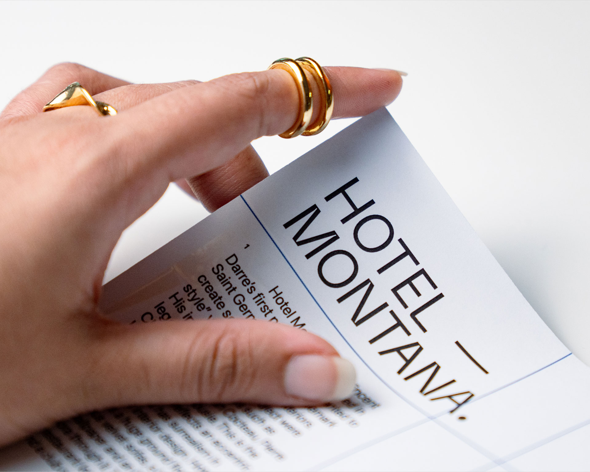 A hand with gold rings gently turns a page of a brochure or magazine showing the bold, uppercase text "HOTEL MONTANA," suggesting travel or hospitality themes.