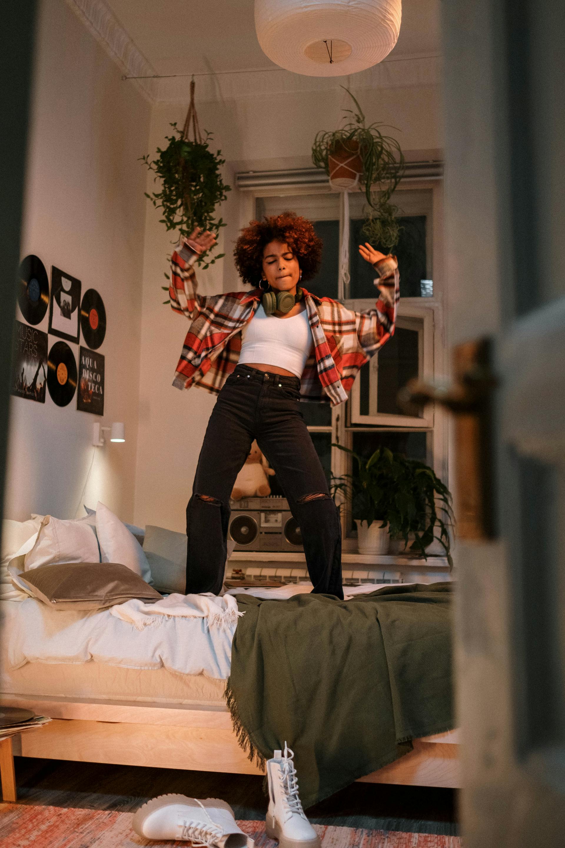 A young woman in a white crop top and black jeans dancing on her bed in a room decorated with hanging plants and vinyl records.
