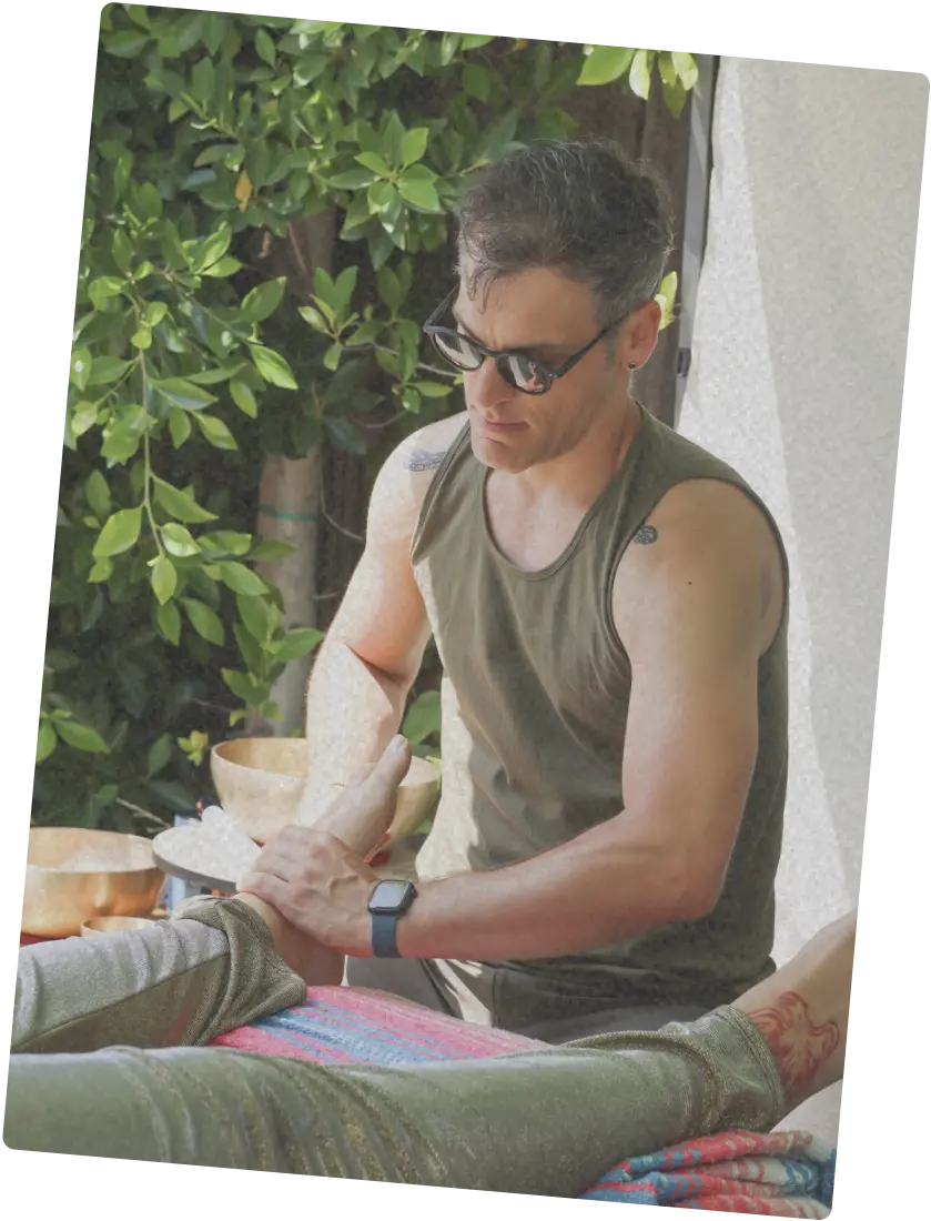 Person wearing sunglasses giving bodywork outdoors, seated beside a person lying on a blanket.