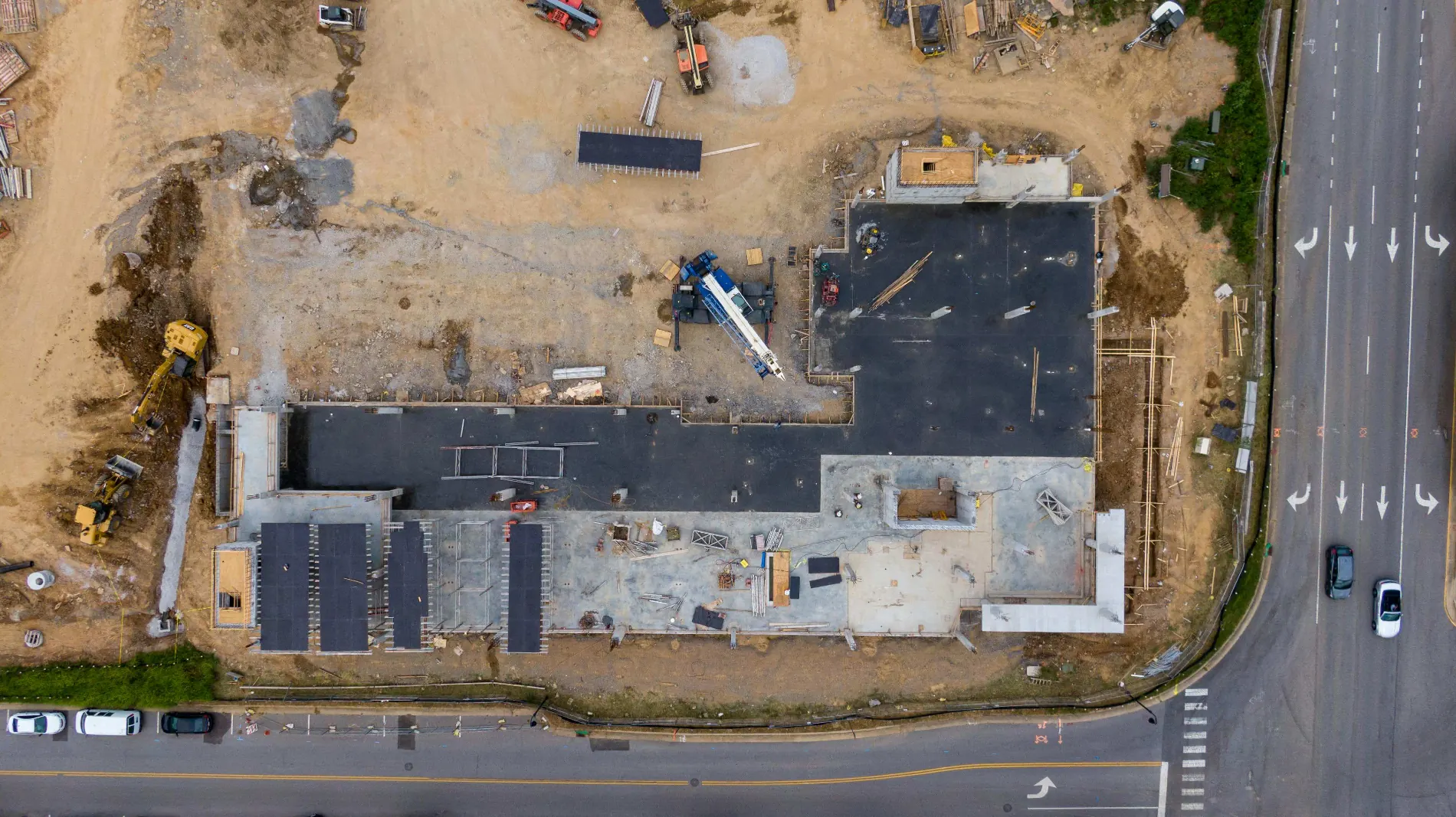 Top-down site progress photo showing building foundation layout, yellow excavators, and adjacent road traffic