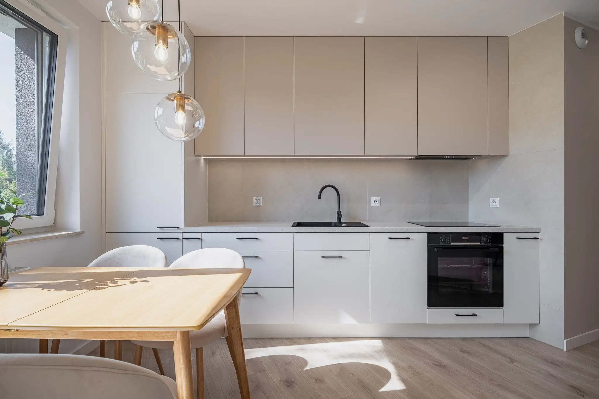 Jasna kuchnia na wymiar z beżowo-białymi frontami, drewnianym stołem i czarnym sprzętem AGD.