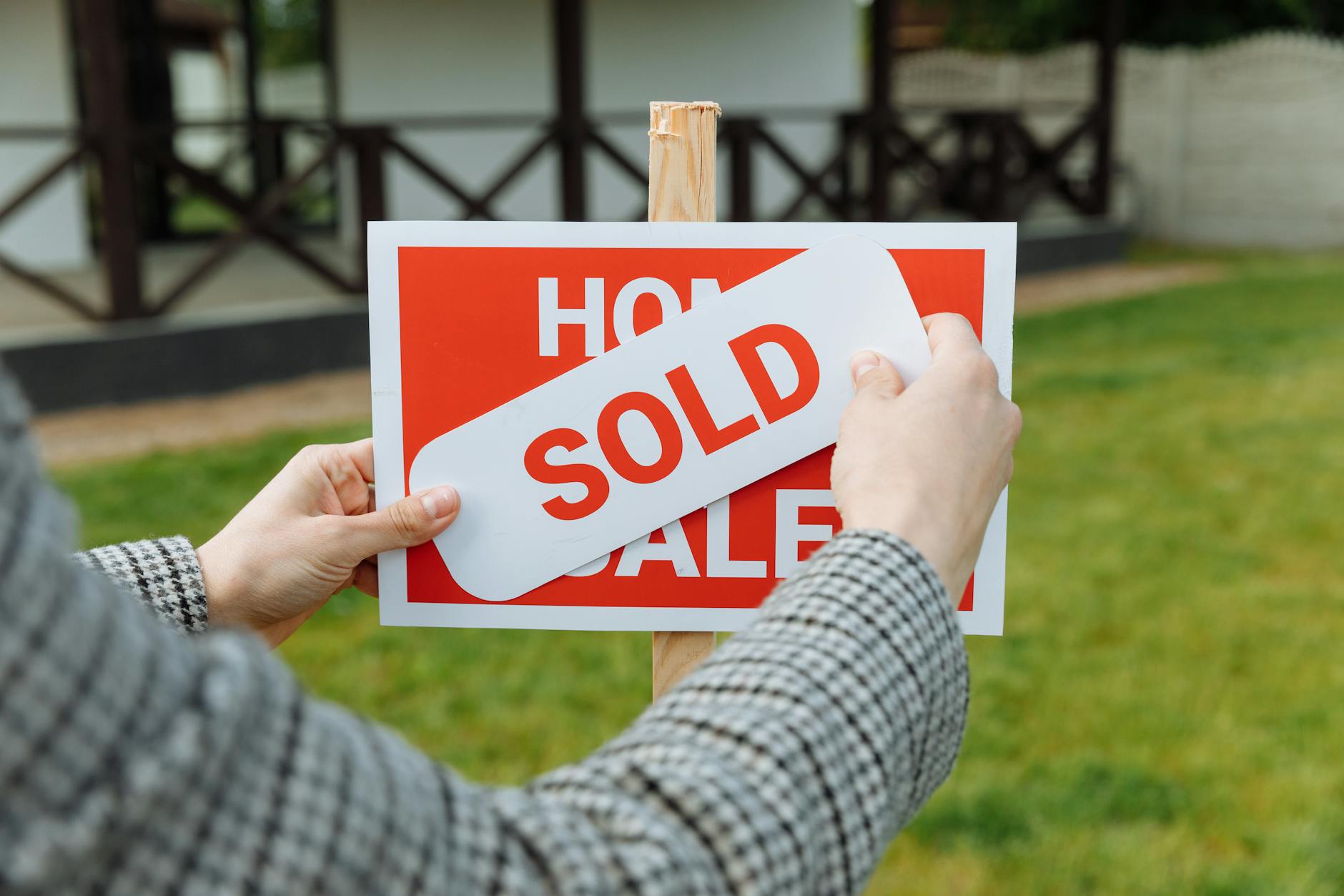 Hands attaching a white Sold sticker over a red Home For Sale sign in a garden setting