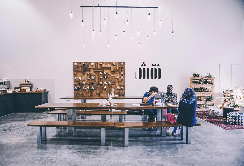 Three people are seated at a long wooden table in a bright, minimalist room. The table is flanked by benches and has a few items on it, including a pink bag. To the left, a dark counter holds various items, and a wooden wall behind it displays a collage of objects. To the right, a shelving unit holds plants and decorative items. A black metal bar with hanging white bulbs runs across the ceiling. The floor is light gray concrete, and the walls are white.