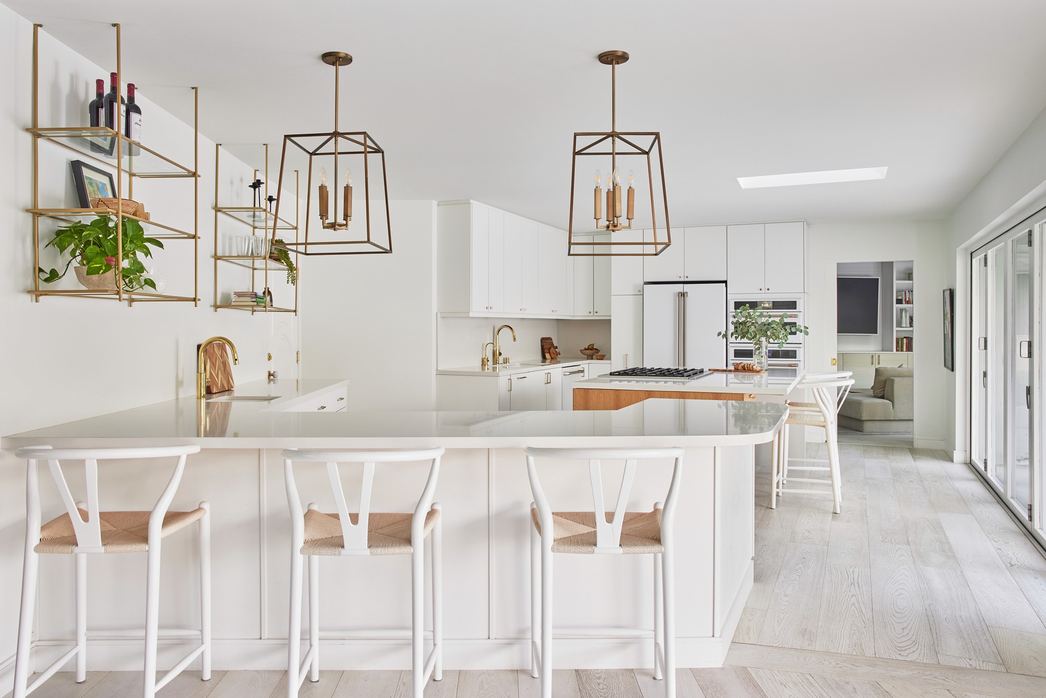 Fresh open concept kitchen with white cabinets, paneled refrigerator, open shelving, bright white quartz countertops, white bar stools, warm wood accents, and light wood flooring by Jubilee Interiors in Los Angeles, California