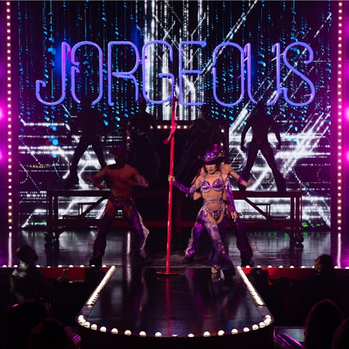 Performer in purple attire and hat dancing on stage with "JORGEIOUS" lit up behind, accompanied by backup dancers in shadow.