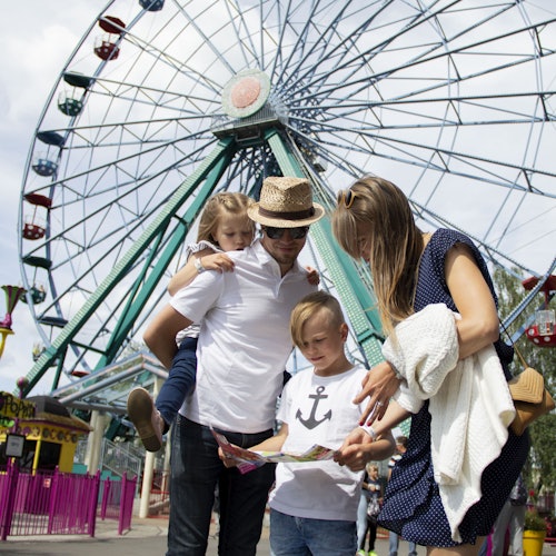A family of four at an amusement park stands in front of a large Ferris wheel, looking at a map.