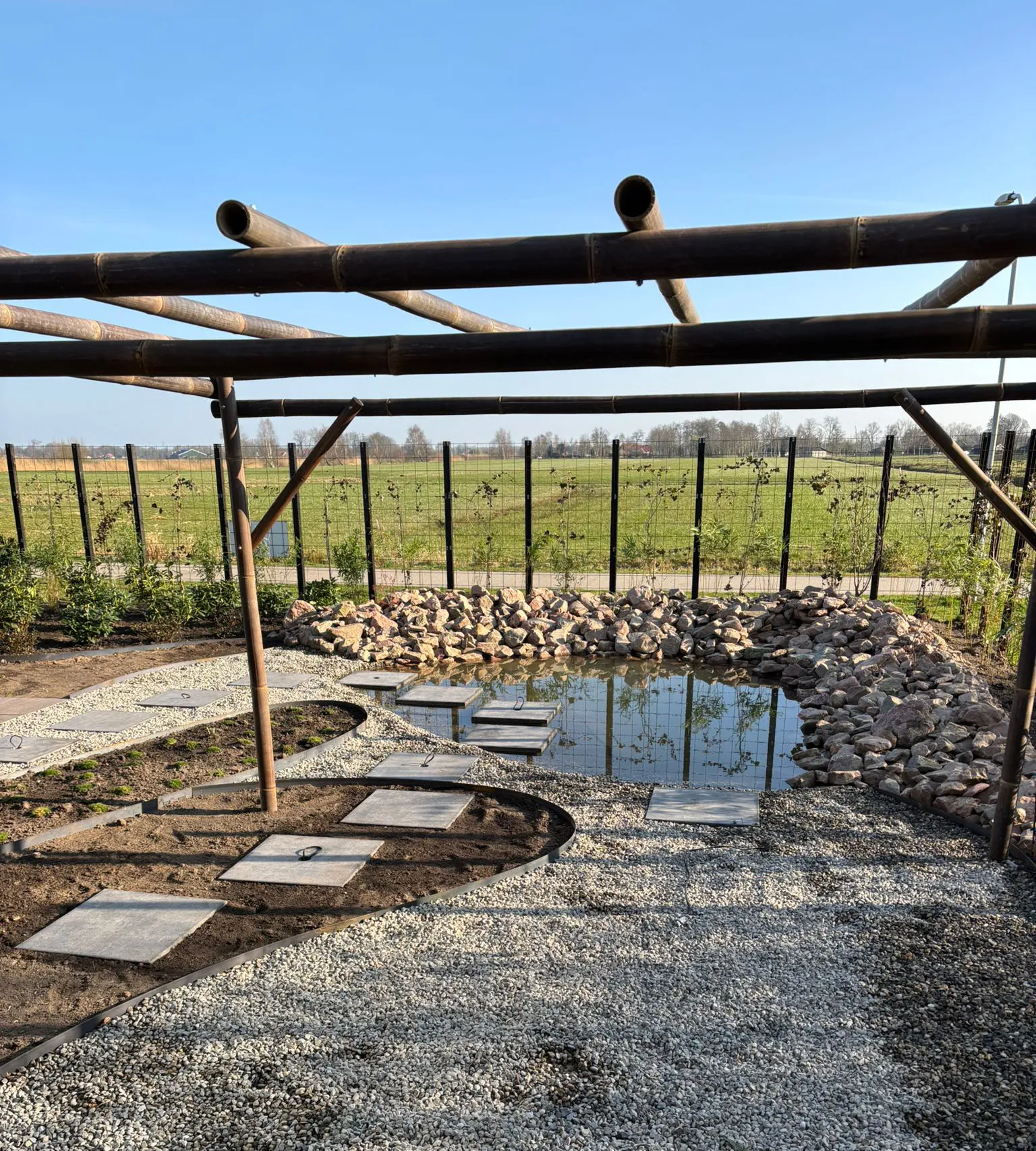 Japanse tuin met vijver omringd door rotsen, grindpad met betontegels en houten pergola, uitzicht op Friese landschap.