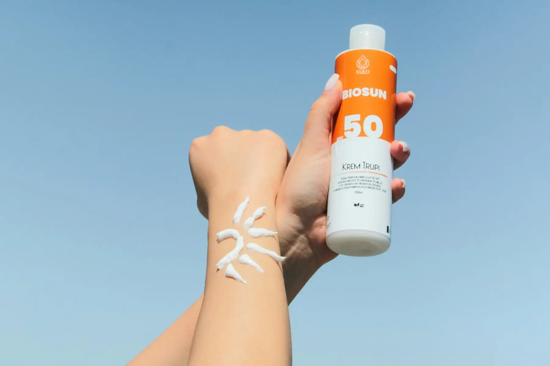 A hand holding an orange SPF 50 bottle against a clear blue sky, sun cream streaked on the forearm