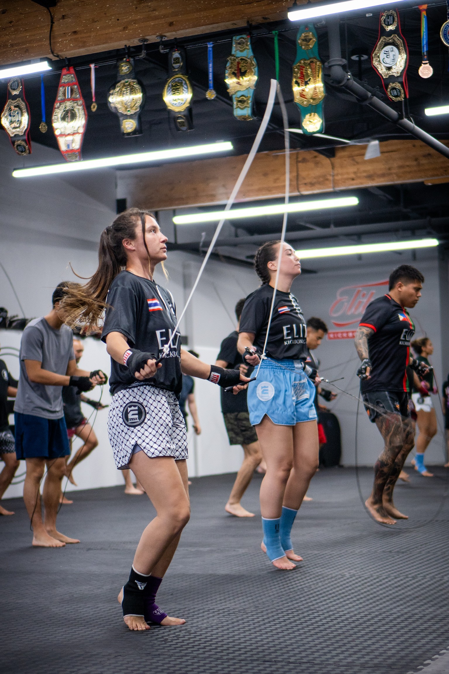 A muscular man smiling while performing a hanging leg raise on gymnastic rings in a bright fitness studio.