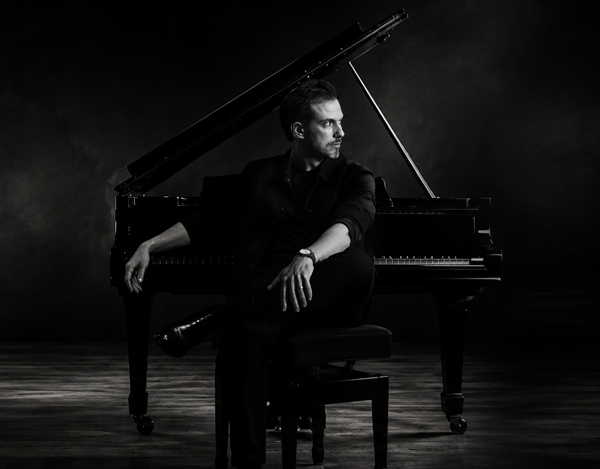 Maximilian Wenzel, German composer, seated at a grand piano in a dark studio setting.