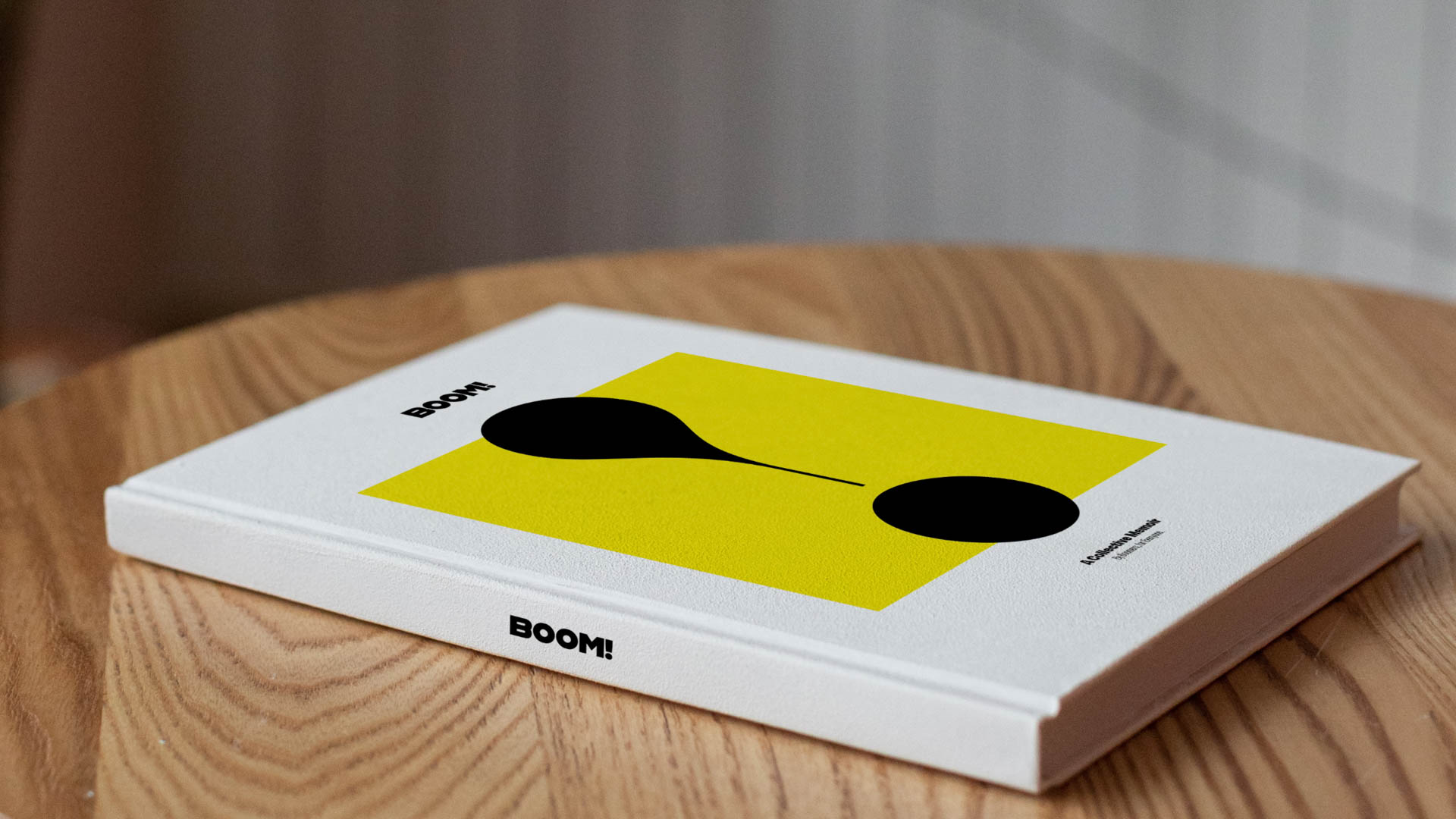 The image shows a white book resting on a light brown wooden surface. The book's cover features a bright yellow square with two black, teardrop-shaped designs. The word "BOOM!" is printed in black text on the cover. The spine of the book also bears the word "BOOM!". In the background, a light-colored wall is visible.