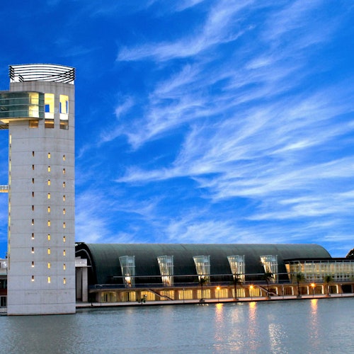 一座現代建築，擁有高聳的觀景塔，矗立在平靜的水面旁，在晴朗的，部分多雲的藍天下。