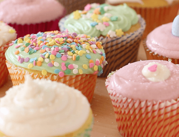 The image shows a wooden table with at least seven cupcakes arranged on it. The cupcakes have colorful frosting in various shades, including pink, green, white, and light blue. Some cupcakes are topped with rainbow sprinkles, while others have swirls of frosting. The cupcakes are in red and white striped paper liners, and some are nestled in multicolored paper cups. The cupcakes are arranged in a somewhat overlapping manner, with some positioned closer to the viewer and others further away. The wooden table provides a natural and warm backdrop for the colorful cupcakes.