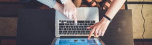 Laptop in the middle of a table with two peoples hands over it