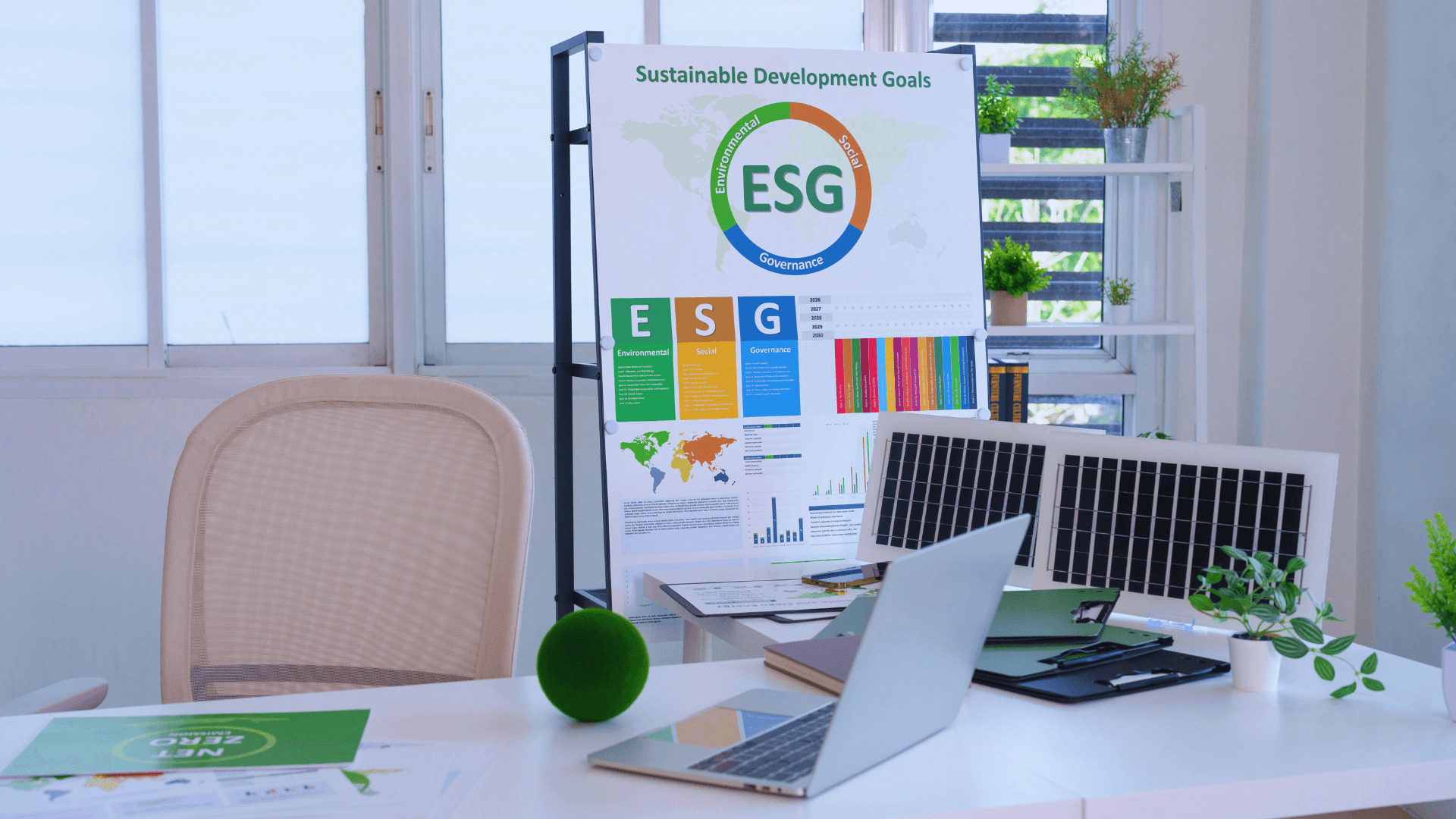 Modern ofis masasında sürdürülebilirlik raporu hazırlığı: Laptop, güneş paneli maketi, 'Sürdürülebilir Kalkınma Amaçları' ve 'ESG' posteri ile 'Net Zero' baskısı.
