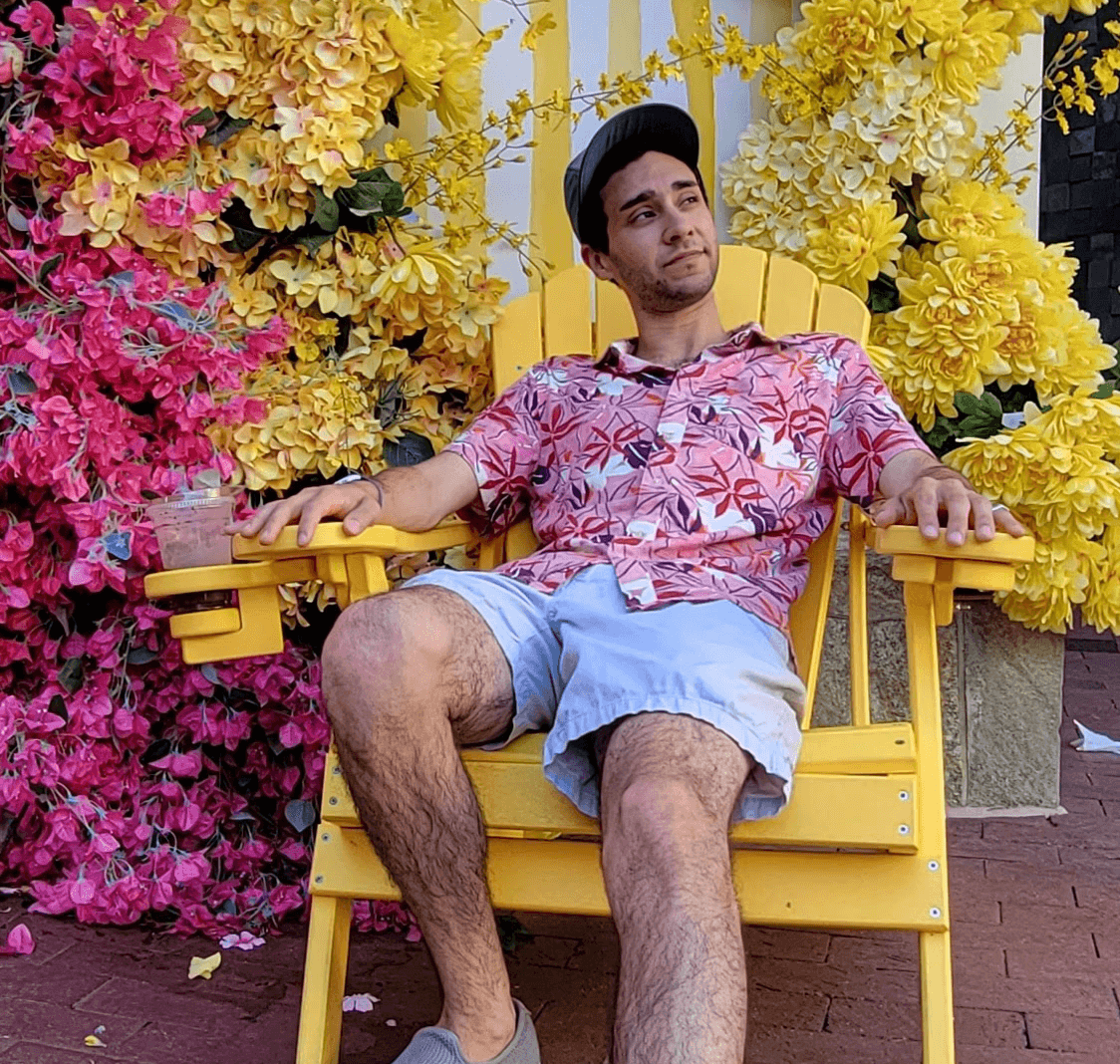 A photo of a man sitting in a wooden chair outside; in the background and multicolored flowers