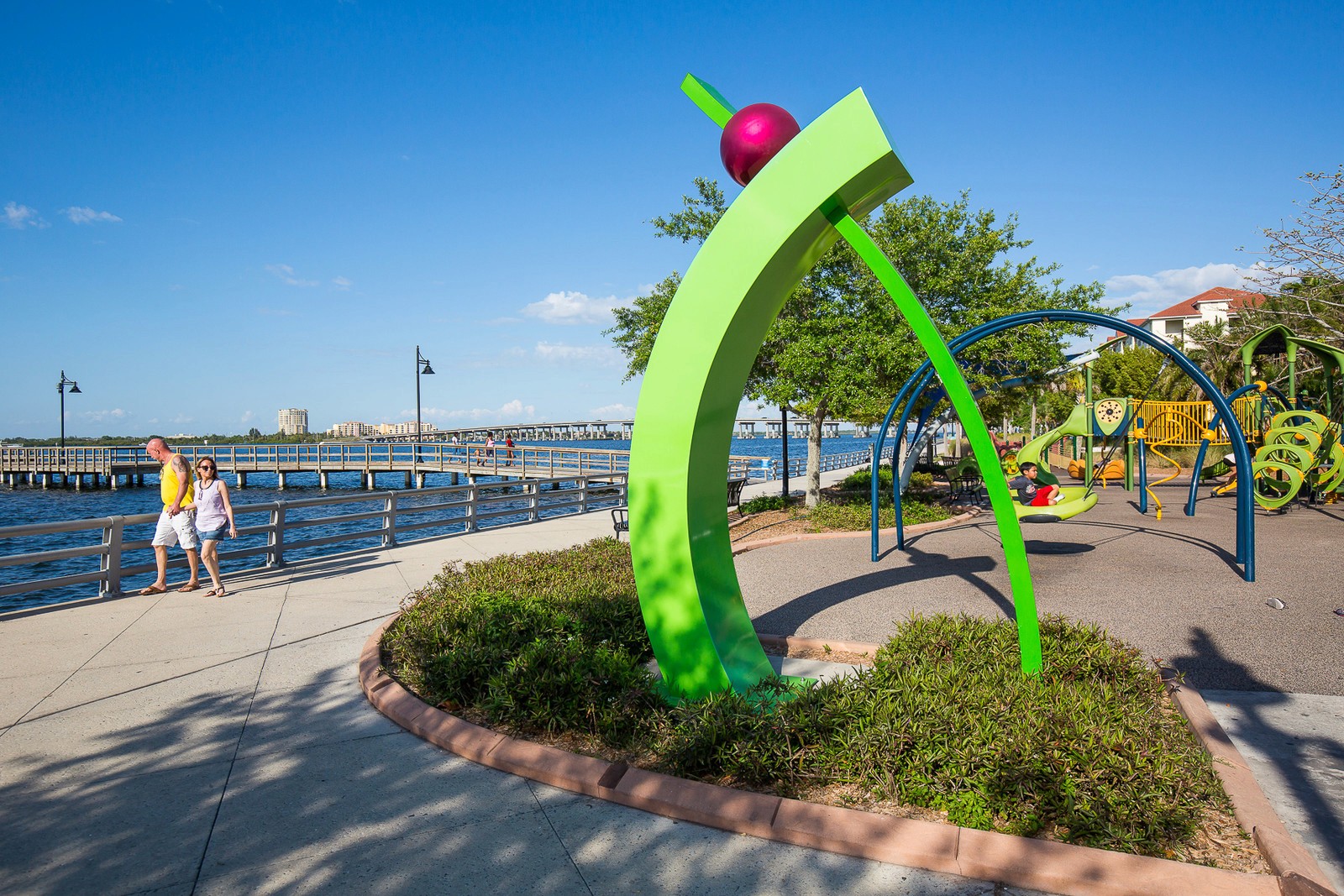 Bradenton Riverwalk waterfront park with a bright green sculpture, a couple strolling the boardwalk, and a children's playground