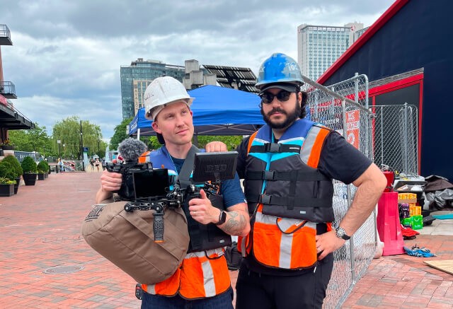 Two men wearing hard hats, safety vests, and life jackets stand outdoors, looking at a camera monitor with construction materials and city buildings in the background—capturing the energy of on-site media production.