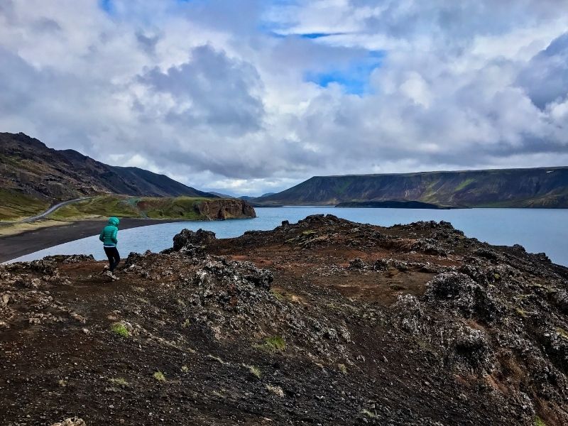 View of Kleifarvatn lake in Iceland with rugged volcanic terrain in the foreground, showing dark, sandy and glassy hyaloclastite layers formed by subglacial eruptions, with mountains and cloudy sky in the background.