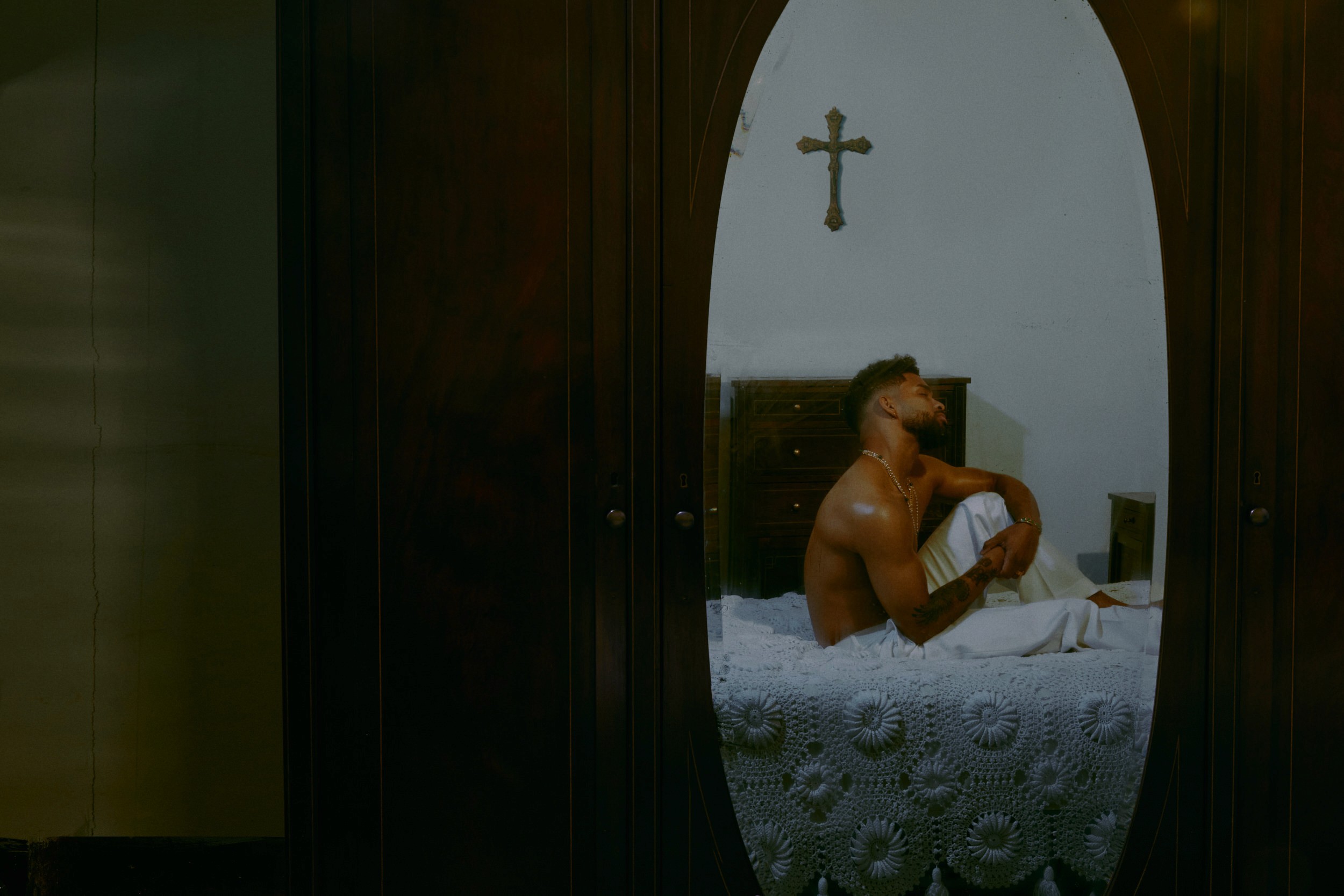 A man sits on a bed viewed through an oval mirror, his back slightly turned, a wooden cross mounted on the wall above him