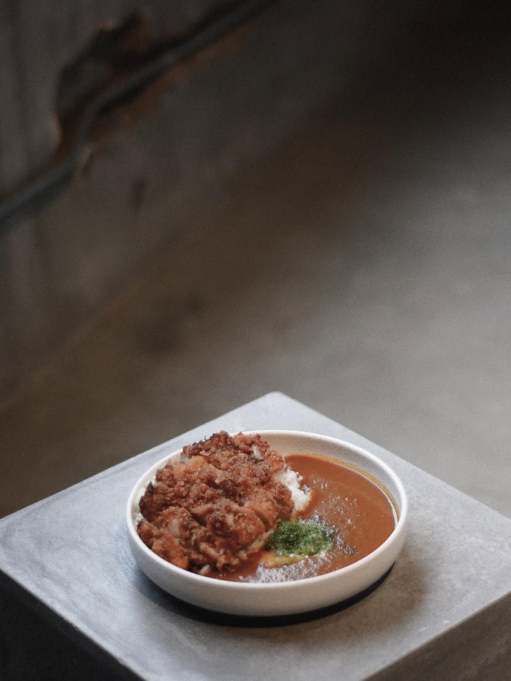 A bowl of curry served with rice and fried chicken, garnished with green sauce, placed on a gray surface.