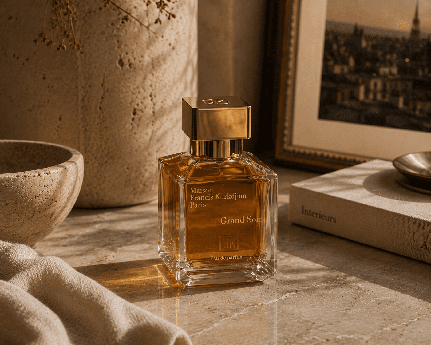 A square clear glass bottle of Maison Francis Kurkdjian Grand Soir Eau de Parfum filled with warm amber liquid, featuring metallic gold lettering and a reflective gold cubic cap. The perfume stands on a light beige stone surface in warm afternoon sunlight next to a stone bowl, a large ceramic vase, draped linen fabric, a book titled "Interiors", and a framed picture showing a cityscape.
