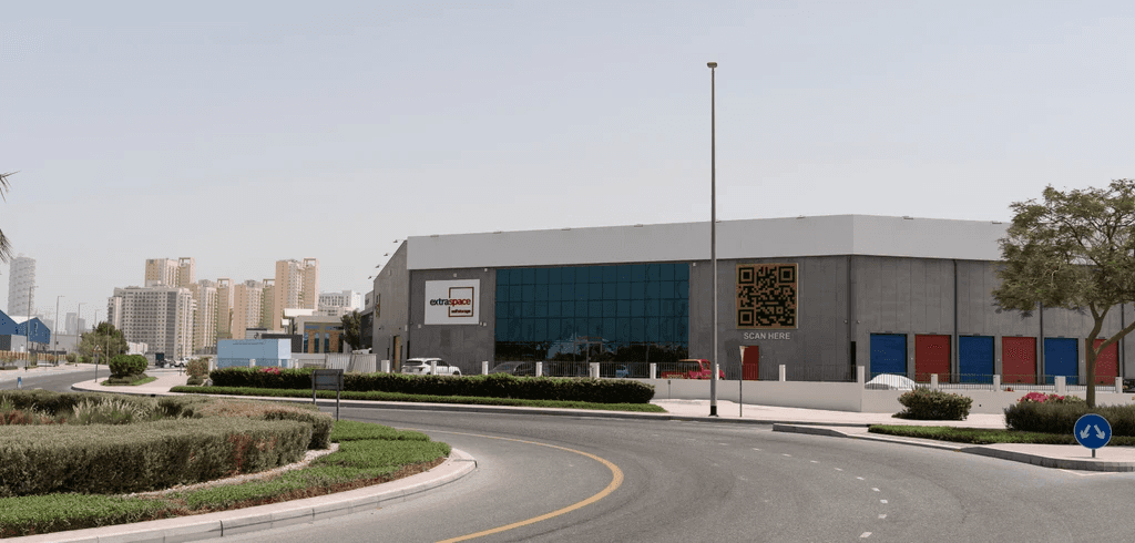 A modern warehouse with a glass facade and large Extra Space sign on the wall is adjacent to a curved road. High-rise buildings are visible in the background.