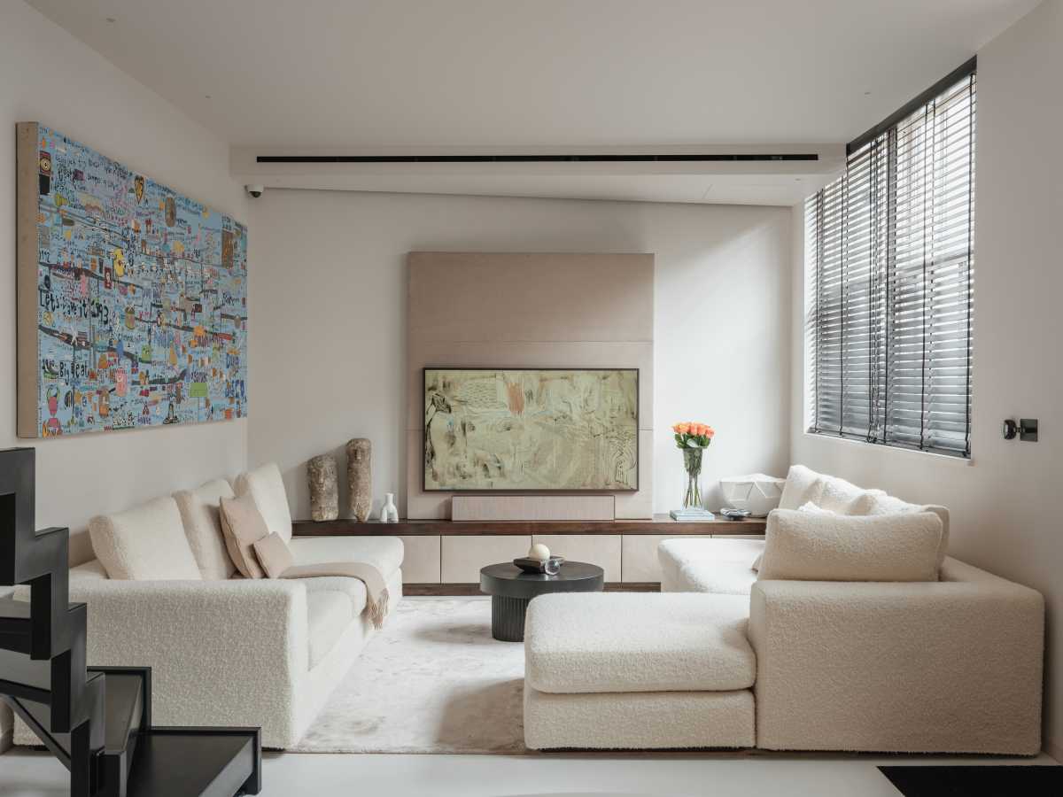 A bright, open-plan modern living room featuring a low-profile cream sectional sofa, a minimalist black coffee table, and custom built-in media cabinetry. Large windows with dark blinds fill the neutral-toned space with natural light, highlighting a seamless architectural flow.