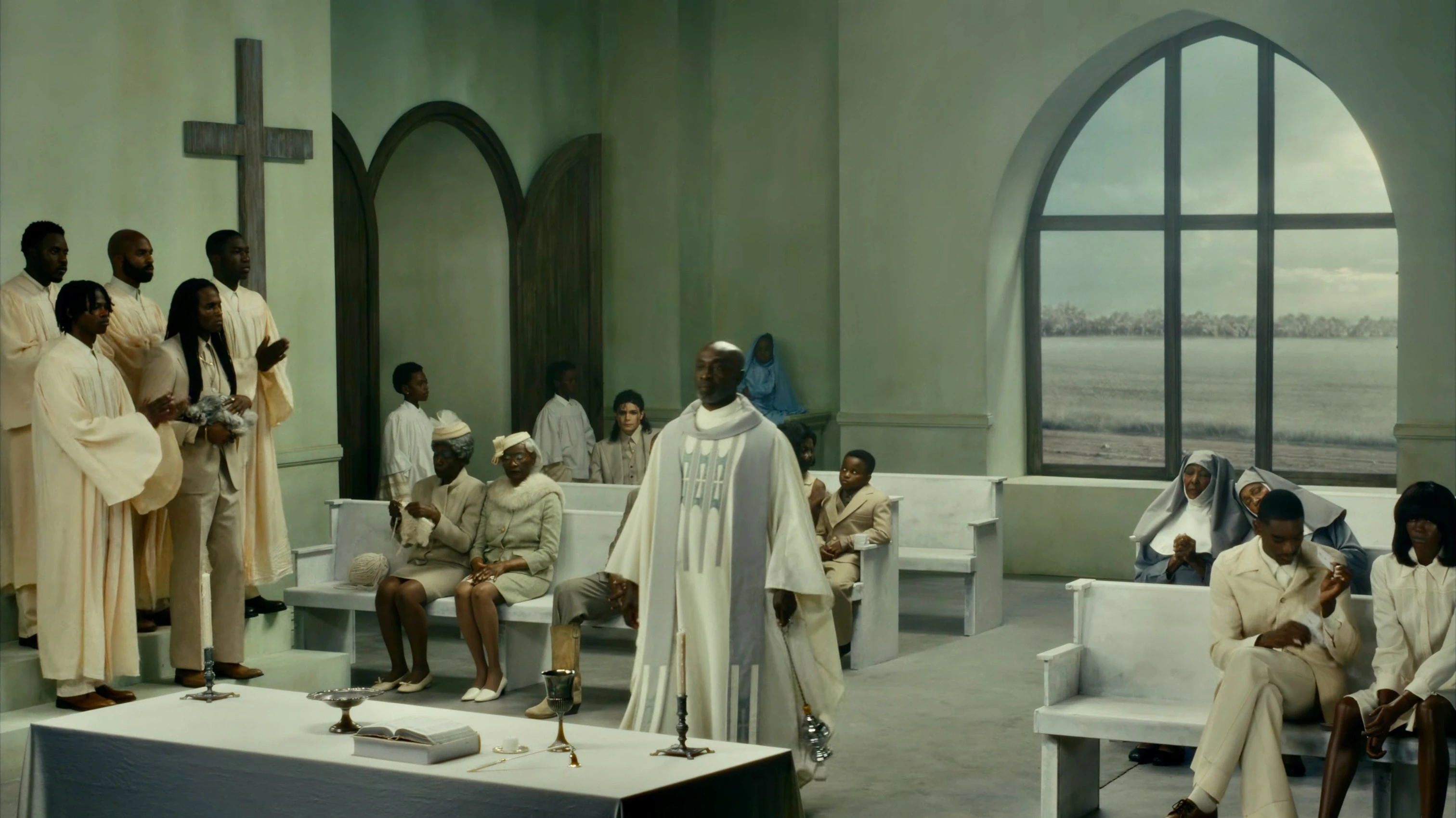 A priest in white vestments stands before a congregation inside a pale green chapel with white pews, a wooden cross on the wall and a large arched window overlooking a flat open landscape