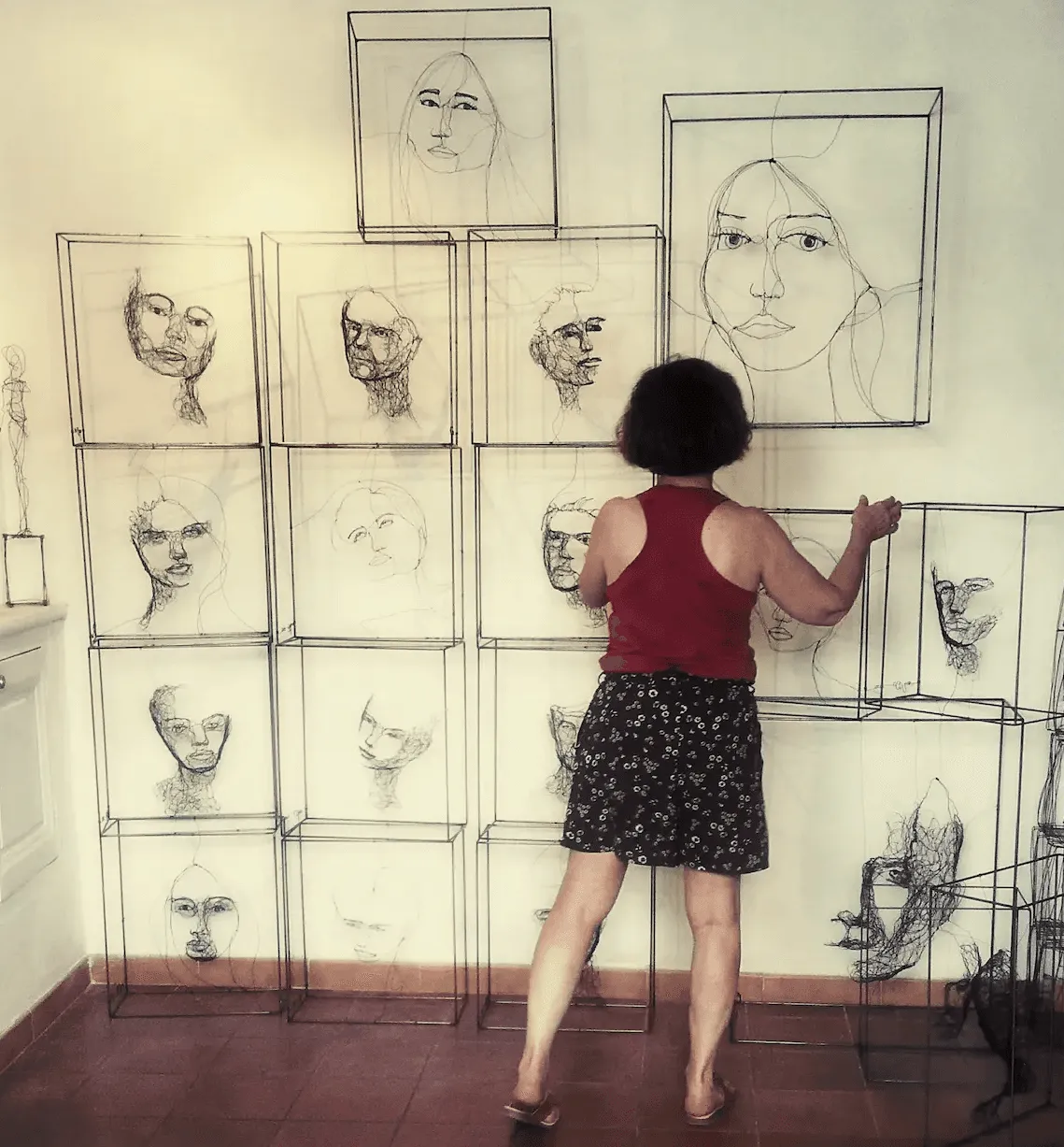 Myriam Louvel de dos fait face à un mur devant lequel une dizaine de ses études de visage en fil de fer sont diposés les unes sur les autres