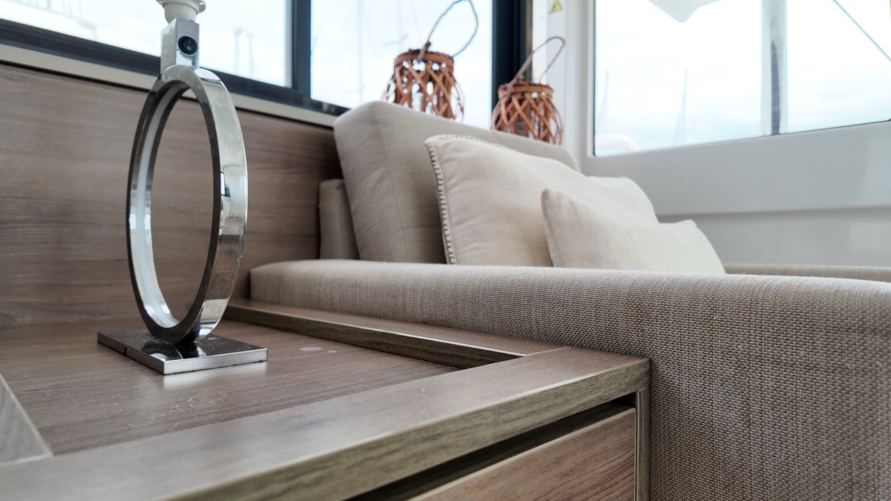 Close-up detail of the Bali 4.4 catamaran main saloon interior showing the linen-upholstered L-shaped sofa with beige cushions, warm grey-oak table surface, chrome ring accent lamp, and wicker lantern decorative accessories on the sill against panoramic black-framed saloon windows