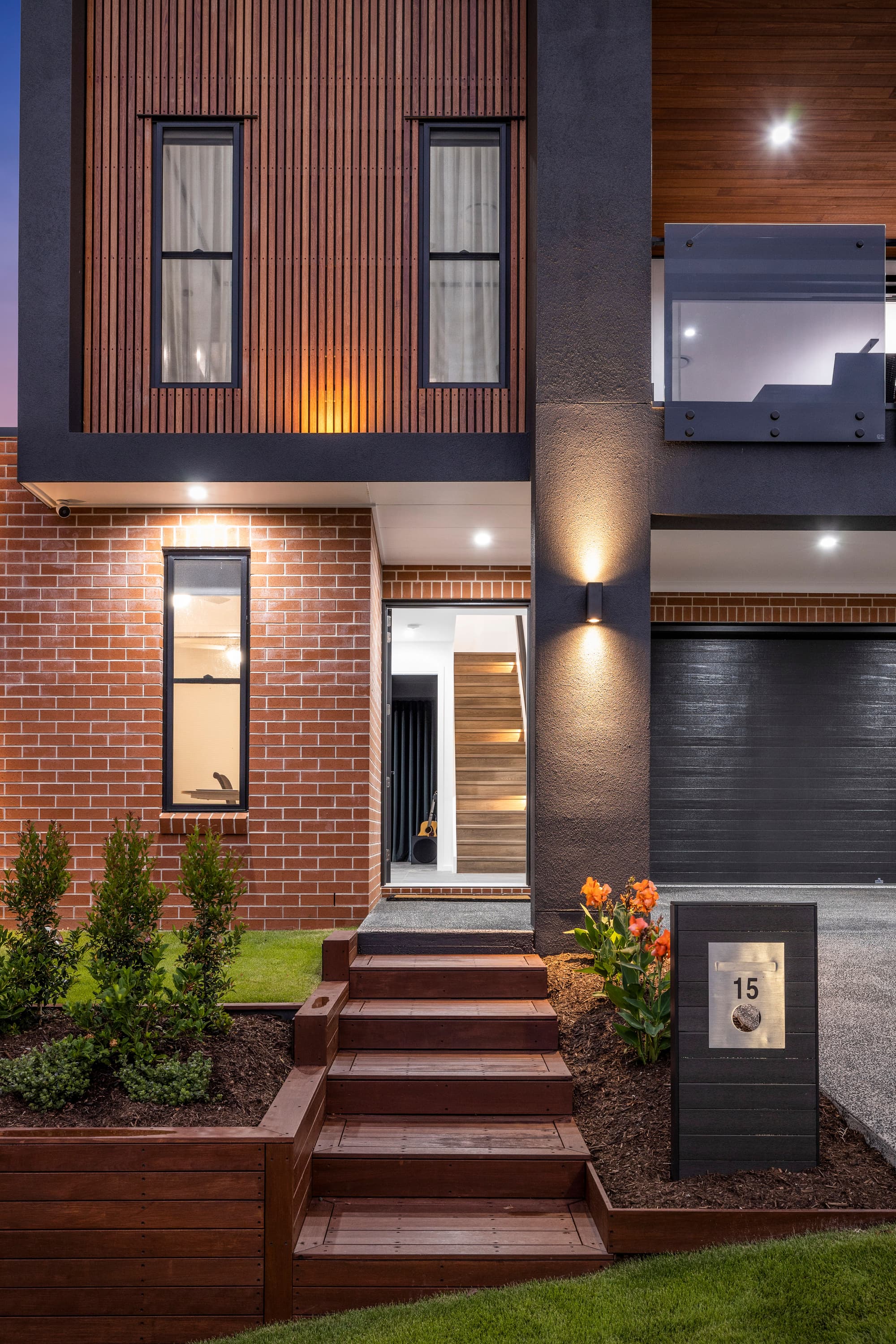 Illuminated entrance of the Stradbroke home with timber steps, red brickwork, vertical timber cladding and landscaped garden beds.