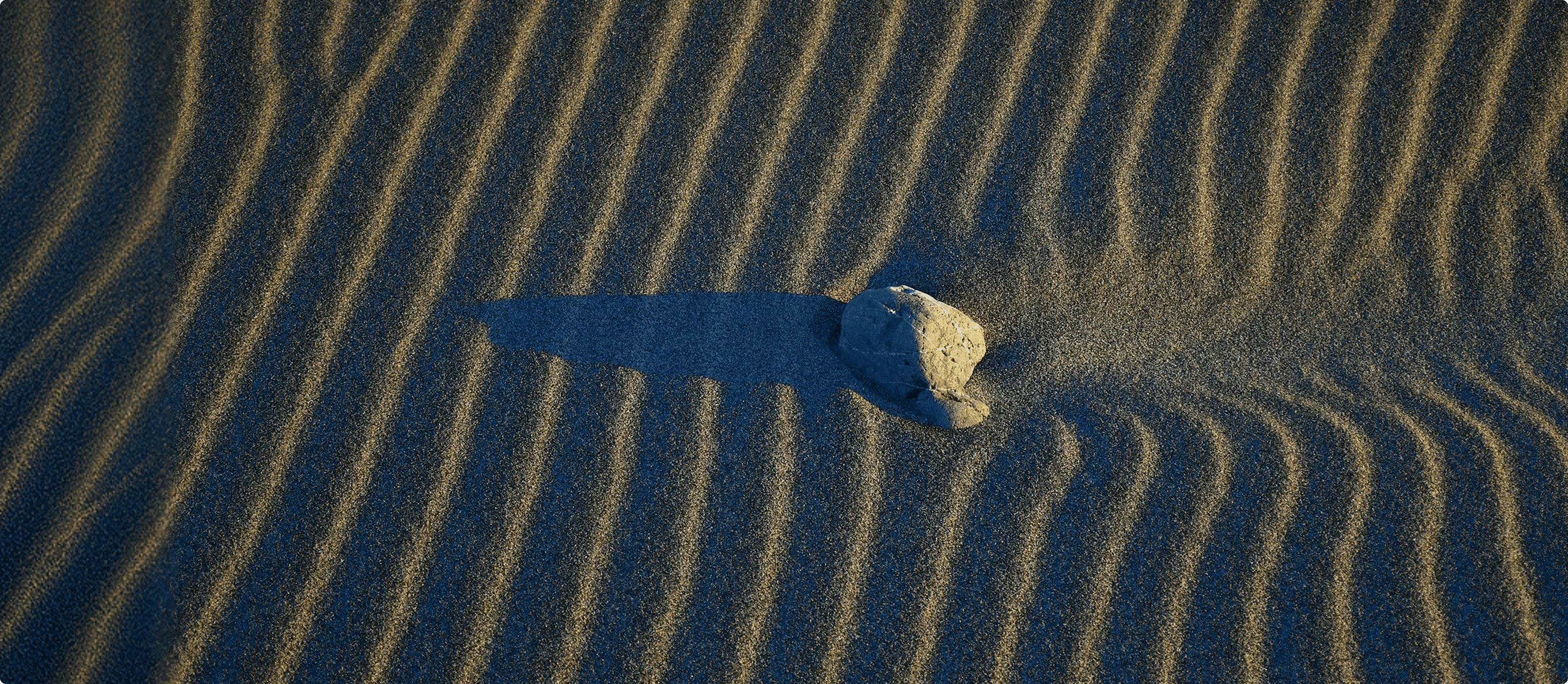  A high-angle shot of a single rock on a sandy beach, casting a long shadow across a rhythmic pattern of wind-rippled sand dunes in the low evening light.