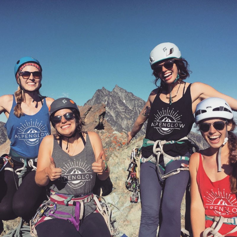 Five women wearing Alpenglow branded tank tops and climbing helmets smile for a photo on a mountain summit.