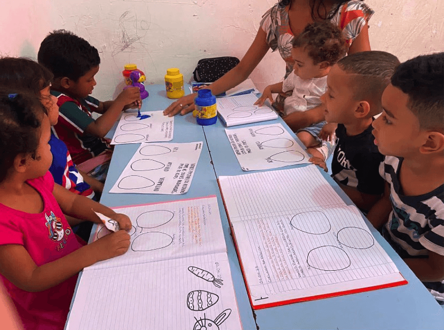 Crianças sentadas ao redor de uma mesa com cadernos de atividades abertas, acompanhadas por uma mulher segurando um bebê.