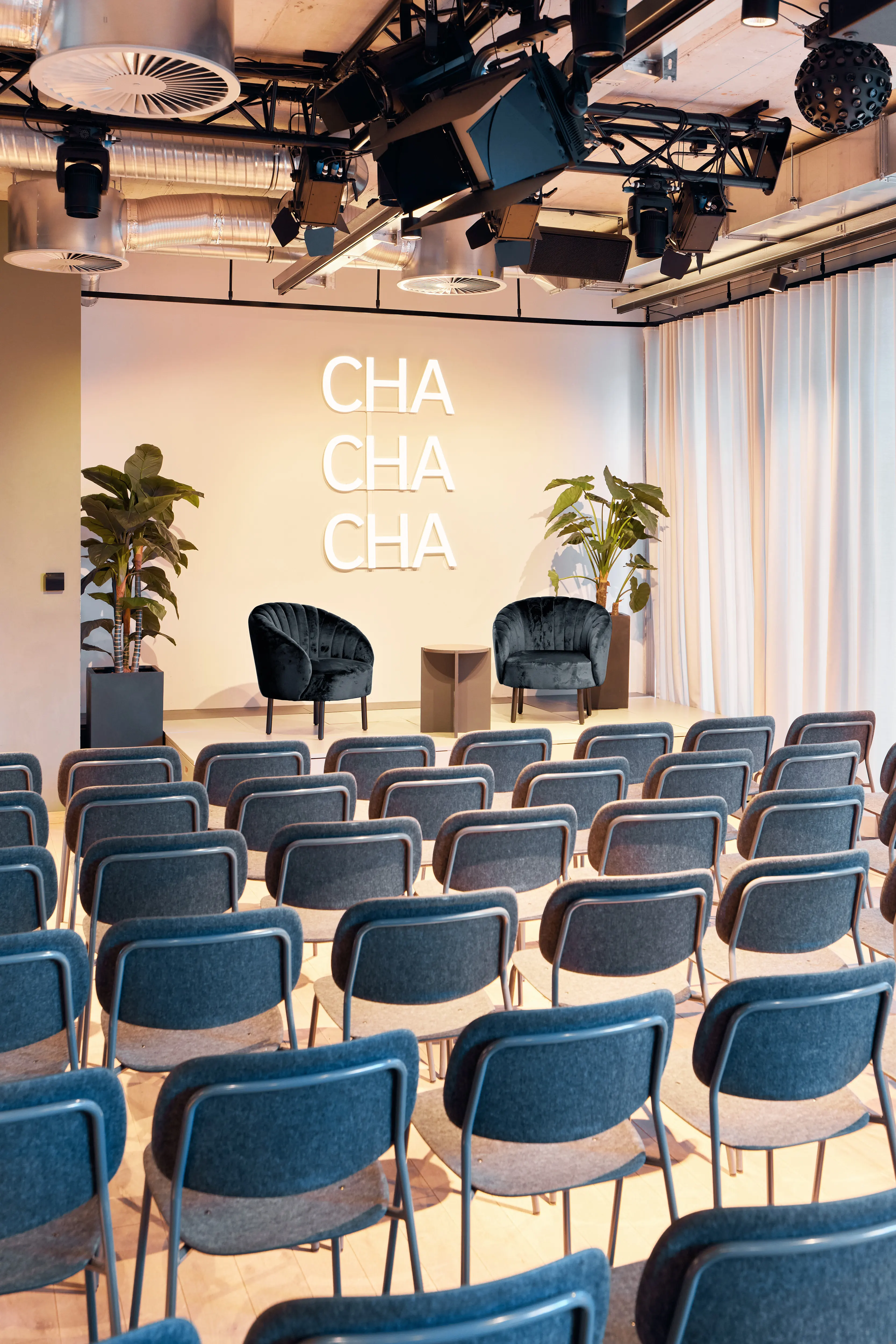 Rows of blue-grey chairs at 381 EVENT SPACE facing a stage setting with two dark green armchairs, plants and CHA CHA CHA lettering on the wall.