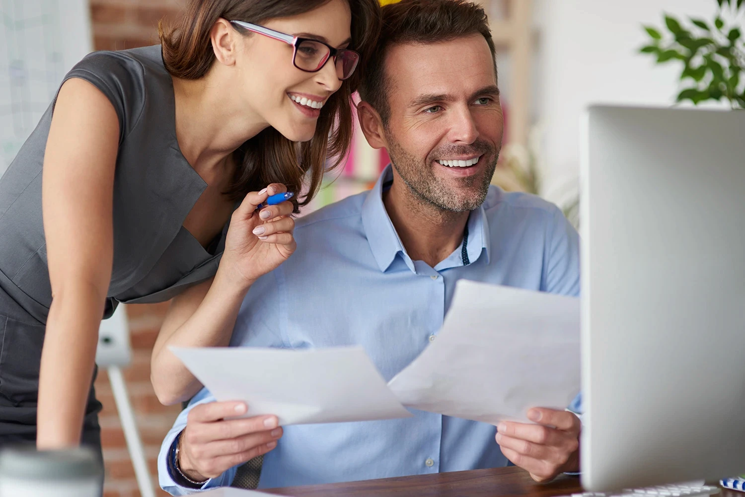 Happy marketing colleagues sitting at a computer, comparing data from paper with information on the screen.