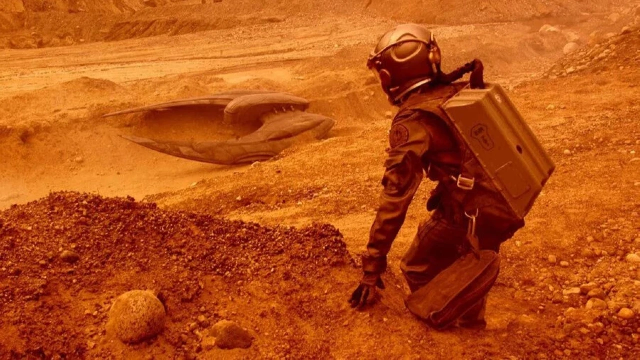 An astronaut in a rugged space suit kneels on the reddish, rocky surface of Mars, observing a mysterious, partially buried alien spaceship in the Martian-like landscape.
