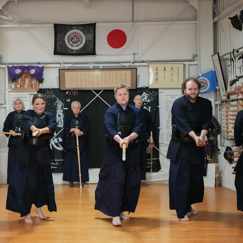 Een groep mensen in traditionele martial arts kleding oefent met houten zwaarden in een dojo, met vlaggen en banieren op de achtergrond.