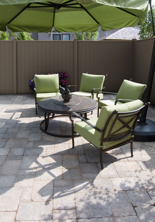 An outdoor stone patio with a dining table, green chairs, and a patio umbrella, bordered by a gray aluminum privacy fence.