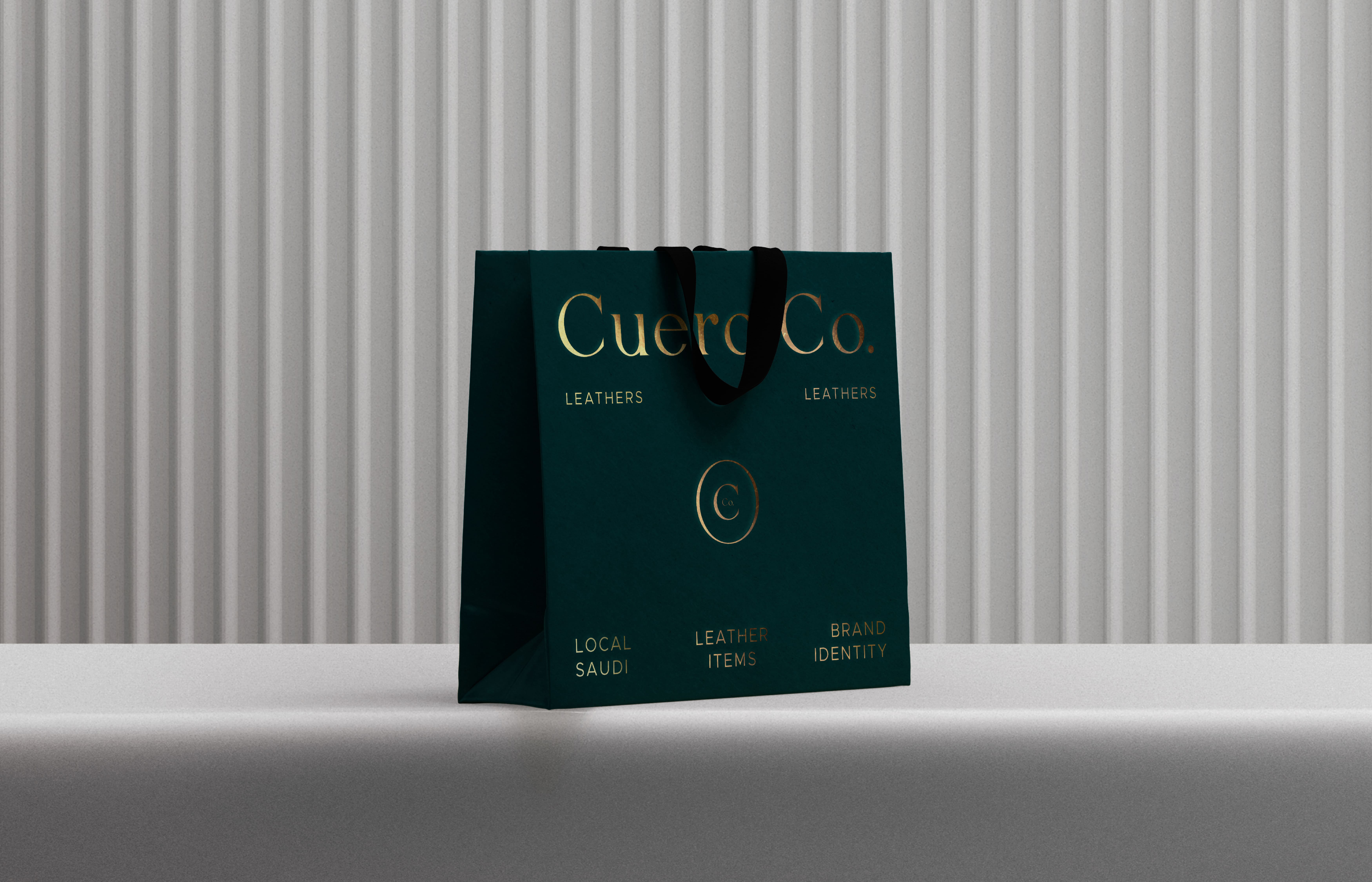 A green shopping bag featuring gold foil lettering that reads Cuero Co. appears against a light gray background with vertical stripes. The bag has black handles and includes additional text: Leathers, Local Saudi, Leather Items, and Brand Identity. The bag is placed on a simple, light-colored surface, illuminated softly to highlight its design.