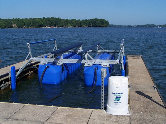 A boat lift with a blue floating mechanism is secured to a dock on a calm lake, surrounded by lush green trees along the distant shoreline under a clear blue sky.