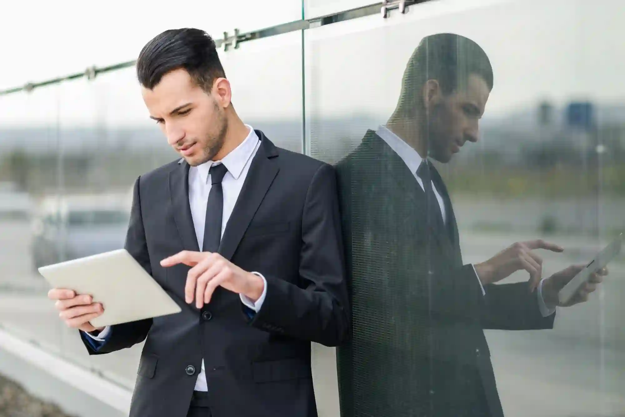 A businessman in a suit uses a digital tablet while leaning against a glass wall in an urban setting.