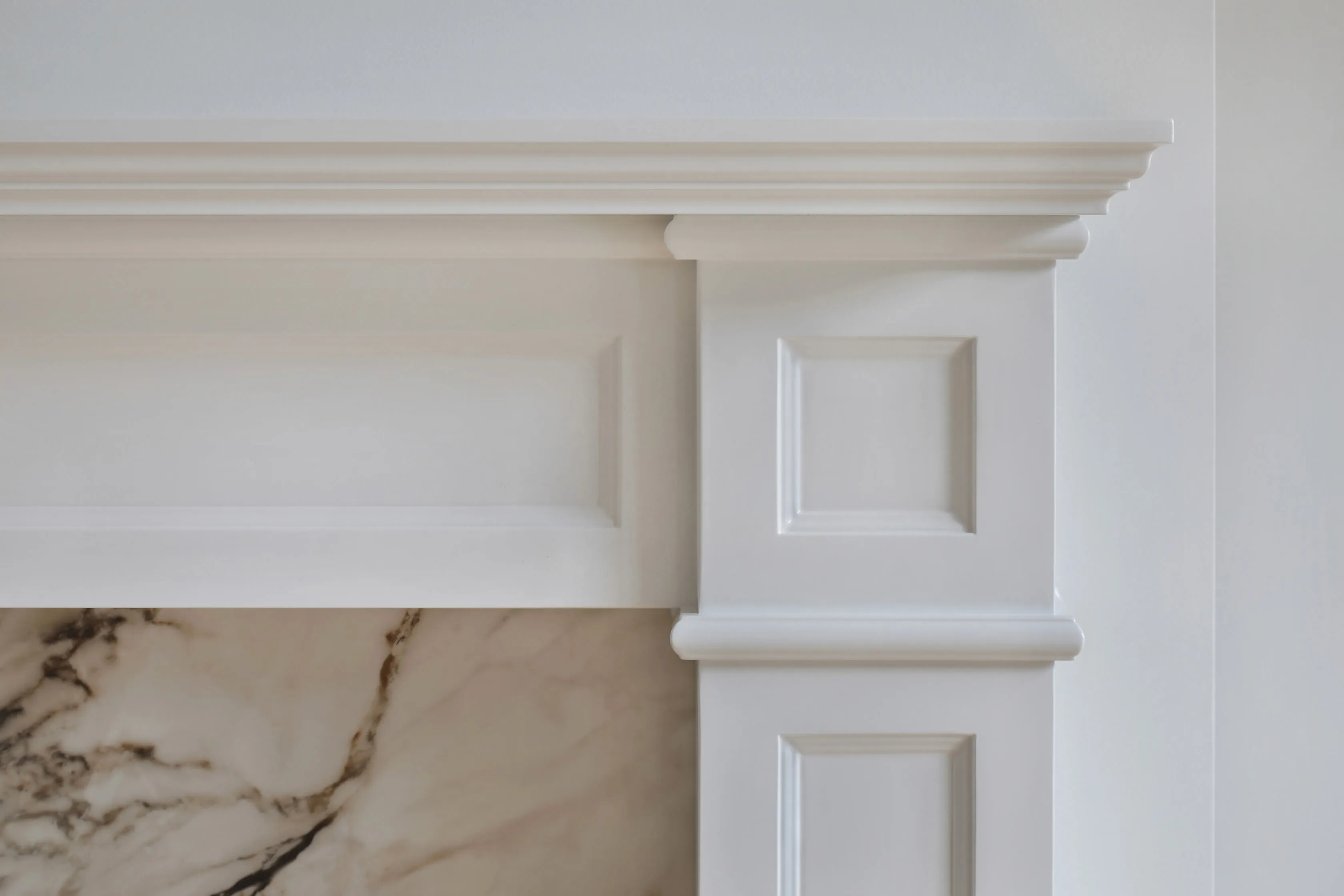Detail of a white classical fireplace mantel with panelled pilasters and moulded cornice against a Calacatta marble surround