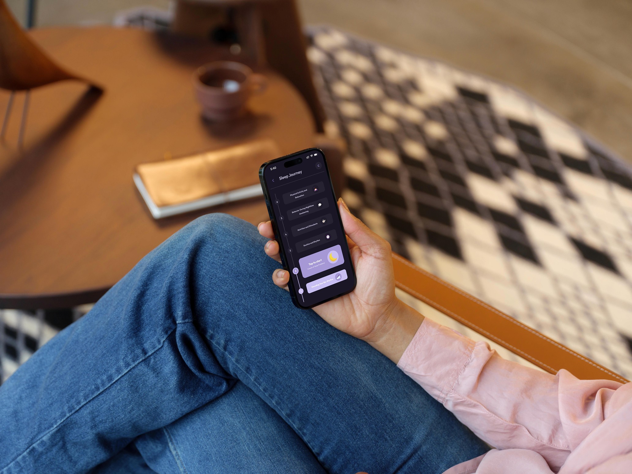 Lifestyle photography of a person sitting comfortably with crossed legs, wearing blue jeans and a pink blouse. They are holding a smartphone displaying the Vialog "Sleep Journey" interface with its timeline and moon icon. The background features a blurred wooden coffee table with a notebook and a patterned rug, creating a cozy atmosphere.