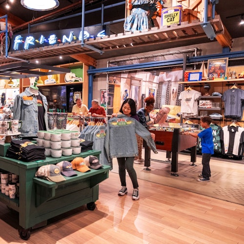 A person holds a "Friends" themed sweatshirt in a store filled with "Friends" merchandise. Children browse items in the background.