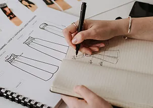 A designers hand using a pen to drawing a pant's flat sketch. There is a book open with few flat sketchs of a pants underneath 
